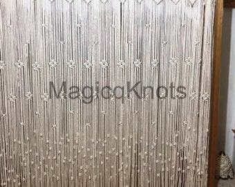 May include: Cream-colored macrame curtain with vertical strands and decorative knots. The text "MagicoKnots" is visible on the curtain. The curtain is hanging in front of a wooden frame. The overall theme is home decor.