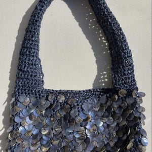 May include: A navy blue crocheted handbag with a matching handle. The bottom of the bag is adorned with iridescent, circular sequins. The bag is designed with a simple, yet elegant style.