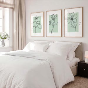 May include: A bedroom scene featuring a bed with white bedding and three framed floral art prints. The prints have a minimalist design with black line drawings on a light green background. A window with white curtains is visible.