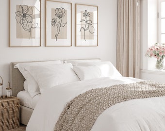 May include: A white bed with a white duvet cover and a beige knitted throw blanket. The bed has a beige headboard and two white pillows. There are three framed black and white line drawings of flowers on the wall behind the bed. A vase of pink flowers sits on a windowsill to the right of the bed.