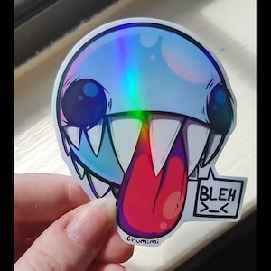 May include: A holographic circular sticker featuring a round, light blue creature with large dark eyes, sharp white teeth, and a long red tongue sticking out. A small white speech bubble contains the black text BLEH >_< and the word Chumimi.