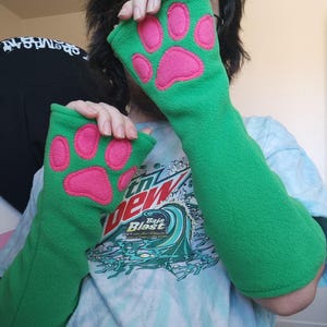 May include: A pair of bright green fleece fingerless gloves featuring vibrant pink paw pad appliqués stitched onto the back of each hand. The gloves extend past the wrists, providing full forearm coverage with a soft, textured fabric finish.