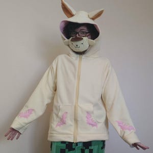May include: Cream-colored zip-up hoodie with a hood featuring bunny ears and a plush face mask. The hoodie has pink bat-shaped appliqués on the sleeves and pockets. The person is wearing glasses and green pixelated pants.