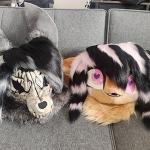 May include: Two furry animal head masks. One is a gray and white wolf with a skeletal face. The other is a black and pink striped cat with pink hearts for eyes.