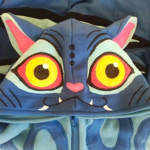 May include: Blue hooded sweatshirt with a cat-like design. The hood features large yellow and red eyes, pink ears, and a small pink nose. The sweatshirt has a zipper closure and is made of a soft, plush material.