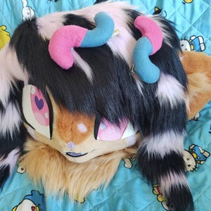 May include: A furry, cartoon-style head with large, expressive eyes and a smiling mouth. The head is primarily tan, with black and white fur accents. Pink and blue horns are attached to the top. The background is a blue blanket with cartoon characters.