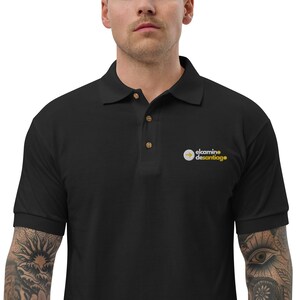 May include: Black polo shirt with a collar and three wooden buttons. The shirt features a small logo on the chest that says "elcaminedesantiage" in yellow and white.