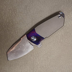 May include: A titanium pocket knife with a damascus steel blade and a blue and purple anodized handle.