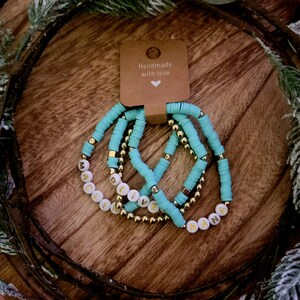 May include: Set of four turquoise beaded bracelets with gold accents. Each bracelet has a white bead with a gold letter spelling out the words "MACI", "OLIVE", "LIVE", and "NANA". The bracelets are handmade with love.