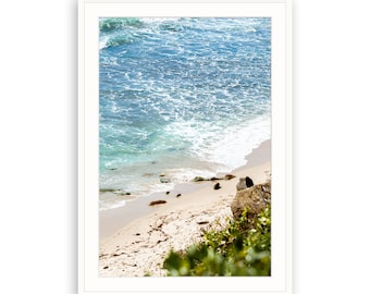 May include: A close-up view of a sandy beach with turquoise water and white waves crashing on the shore. Green foliage is in the foreground.