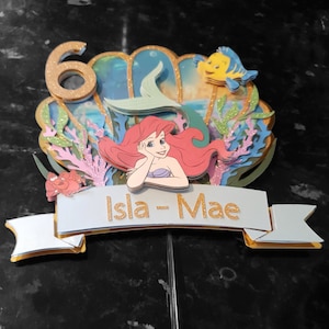 May include: A cake topper featuring Ariel, the Little Mermaid, with a seashell, seaweed, and a yellow fish. The topper also includes a banner with the name "Isla-Mae" and the number "6" in gold glitter.