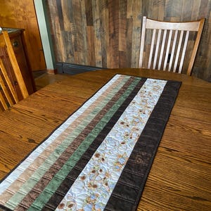 May include: A quilted table runner featuring a central floral design and alternating stripes of brown, green, and beige. The runner is displayed on a wooden table, highlighting its decorative appeal and colour scheme.
