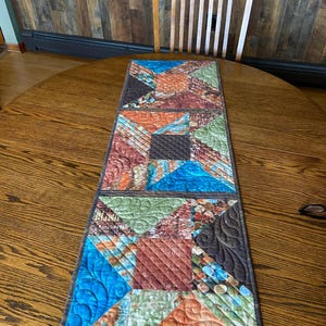 May include: A rectangular quilted table runner with a patchwork design. The runner features squares and triangles in shades of blue, orange, brown, and green. The runner is placed on a round wooden table, with a wooden chair in the background.
