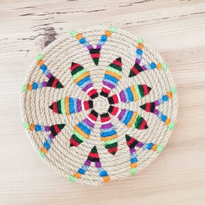 May include: A round, woven jute coaster with a colorful, geometric design. The design features a central circle with radiating lines of different colors, including red, blue, green, yellow, and purple.
