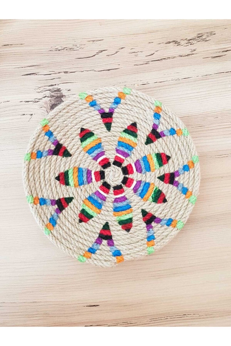 May include: A round, woven jute coaster with a colorful, geometric design. The design features a central circle with radiating lines of different colors, including red, blue, green, yellow, and purple.