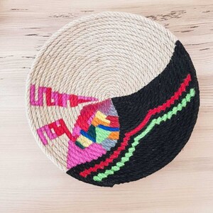 May include: A round woven wall hanging with a black, white, pink, red, green, blue, and yellow geometric design.