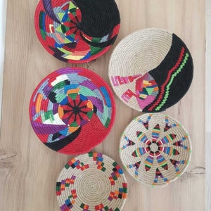 May include: Five round woven wall decor pieces with colorful geometric patterns. The pieces are made of natural fiber rope and are decorated with colorful yarn.
