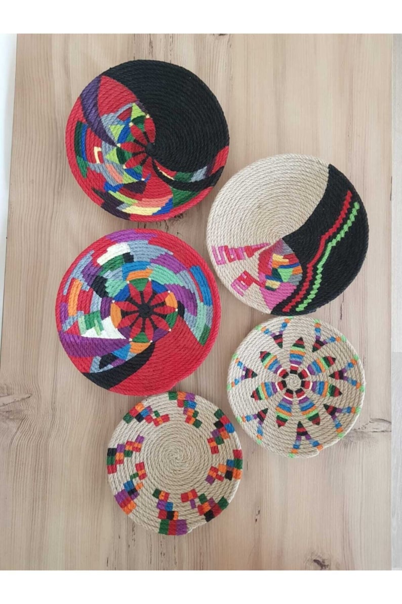 May include: Five round woven wall decor pieces with colorful geometric patterns. The pieces are made of natural fiber rope and are decorated with colorful yarn.