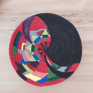 May include: A round, colorful woven wall hanging with a black center. The hanging is made of rope and features a geometric design with red, green, blue, yellow, and purple colors.