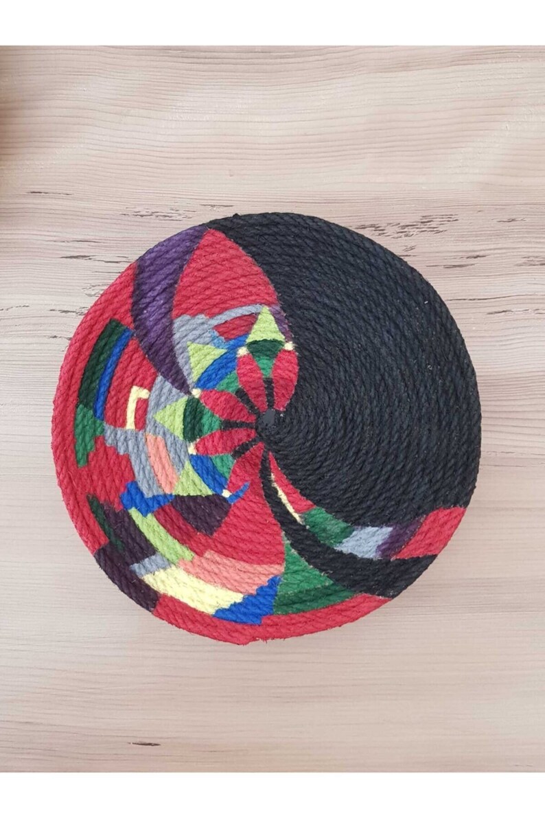May include: A round, colorful woven wall hanging with a black center. The hanging is made of rope and features a geometric design with red, green, blue, yellow, and purple colors.