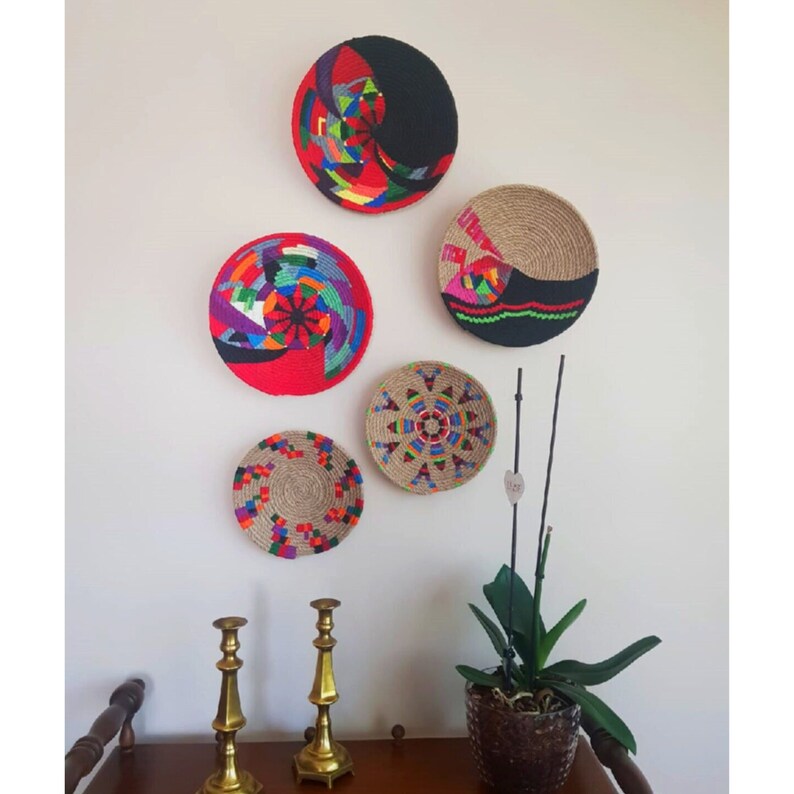 May include: Five colorful woven wall baskets with geometric patterns in red, black, green, blue, and yellow. The baskets are hanging on a white wall with two gold candle holders on a wooden table below.