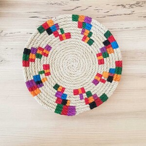 May include: A round, woven coaster with a colorful geometric pattern. The coaster is made of natural fibers and has a rustic, handmade look.