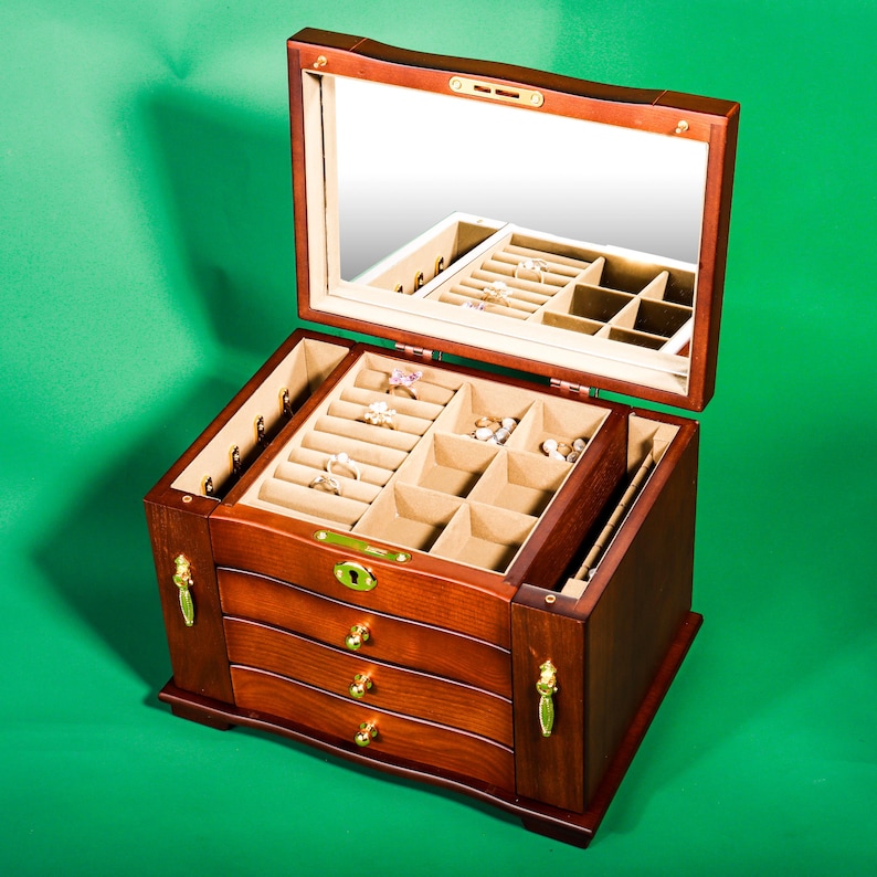 May include: A wooden jewelry box with a mirrored lid and three drawers. The box has a light brown finish and gold-colored accents. The lid is open and the interior is lined with beige felt. The box is filled with various jewelry, including rings, earrings, and necklaces.