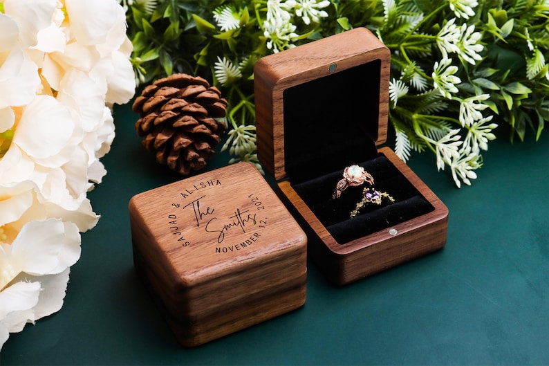 K&ouml;nnte beinhalten: Eine Holzkiste f&uuml;r Ringe mit einer personalisierten Inschrift: "Sajjad & Alisha The Smiths November 12, 2021". Die Box ist ge&ouml;ffnet und ein Ring mit einem gro&szlig;en Edelstein befindet sich darin.