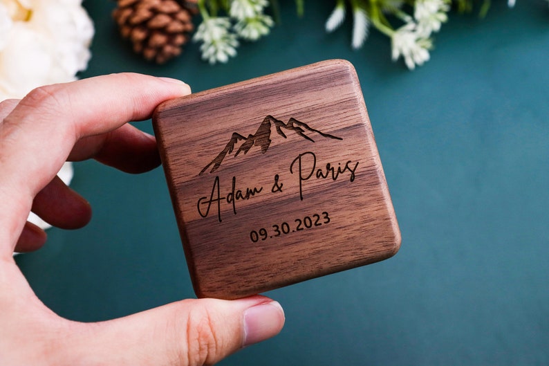 K&ouml;nnte beinhalten: Eine Holzkiste f&uuml;r Ringe mit einem Bergketten-Design und dem eingravierten Text "Adam & Paris" und "09.30.2023" auf der Oberseite.