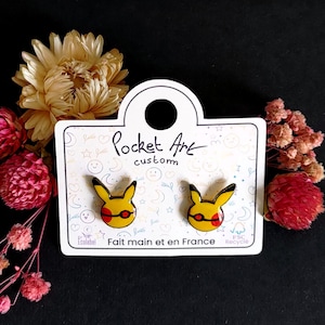 May include: A pair of yellow and red cartoon character earrings on a white card with the text "Pocket Art Custom" and "Fait main et en France".