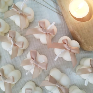 May include: A collection of small, white cloud-shaped candles with a delicate, blush-coloured bow tied around each one. The candles are arranged on a light-coloured surface.