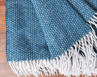 May include: A folded teal throw blanket with a woven pattern and white fringe. The blanket is made of a soft material and is displayed on a wooden surface. The blanket is a square shape.
