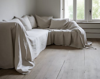 May include: A large, off-white linen sofa with multiple pillows, draped with a linen throw. The sofa is positioned in a corner of a room with a large window behind it.