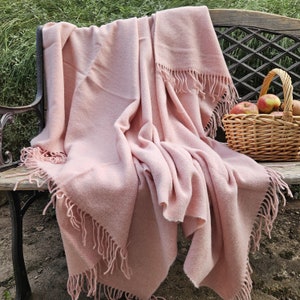 May include: A soft, pink throw blanket with fringe edges draped over a wooden bench. The blanket is made of a lightweight, breathable fabric and is perfect for adding a touch of warmth and style to any room.