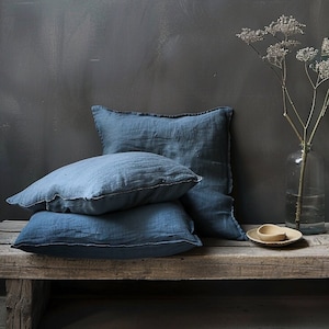 Peut inclure: Trois coussins en lin bleu empilés sur un banc en bois rustique avec un vase de fleurs séchées et un petit bol sur le banc.