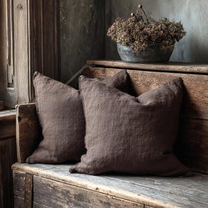 May include: Two brown linen cushions with a textured appearance sit on a rustic wooden bench. The cushions are square and slightly rumpled. A metal container with dried flowers is also on the bench. The scene has a relaxed, natural aesthetic.
