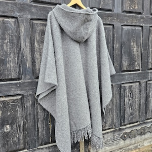 K&ouml;nnte beinhalten: Ein grauer Kapuzenponcho mit Fransen am Saum h&auml;ngt an einem Holzkleiderb&uuml;gel. Der Poncho besteht aus einem dicken, gewebten Material. Der Hintergrund ist eine dunkle Holzt&uuml;r.