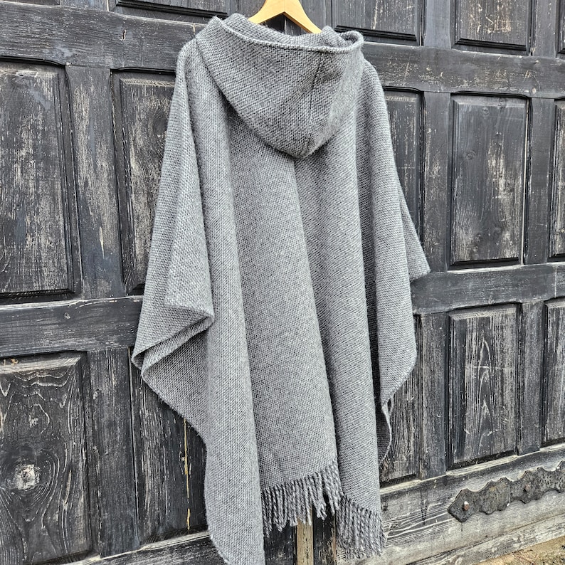 K&ouml;nnte beinhalten: Ein grauer Kapuzenponcho mit Fransen am Saum h&auml;ngt an einem Holzkleiderb&uuml;gel. Der Poncho besteht aus einem dicken, gewebten Material. Der Hintergrund ist eine dunkle Holzt&uuml;r.