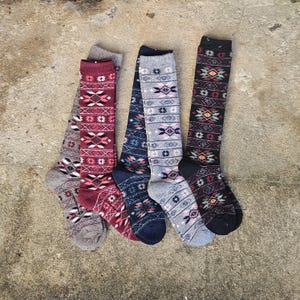 May include: Five pairs of knee-high socks in various colors, including gray, burgundy, navy, and black. Each sock features a geometric pattern in shades of red, white, and orange. The socks are arranged on a textured, neutral-toned surface.