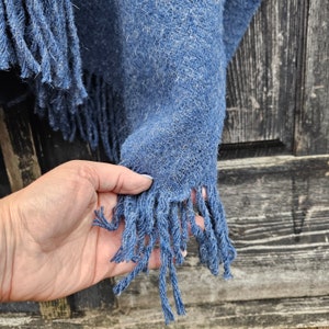 K&ouml;nnte beinhalten: Nahaufnahme eines blauen gewebten Schals mit Fransen. Der Schal hat ein strukturiertes Aussehen und ist einfarbig blau. Die Fransen sind in einem dunkleren Blauton gehalten und h&auml;ngen von der Unterkante des Schals. Eine Hand h&auml;lt den Schal.