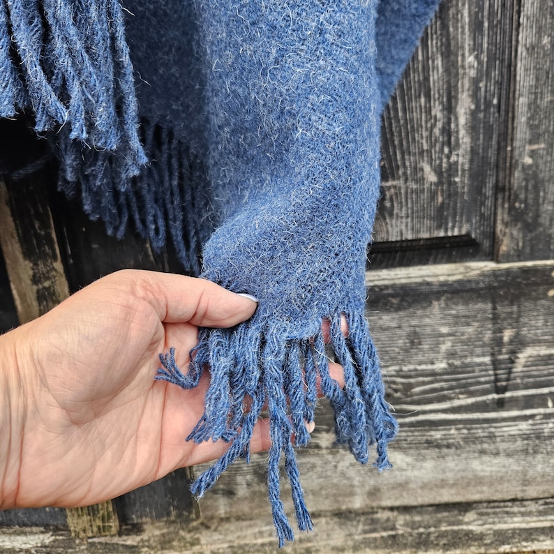 K&ouml;nnte beinhalten: Nahaufnahme eines blauen gewebten Schals mit Fransen. Der Schal hat ein strukturiertes Aussehen und ist einfarbig blau. Die Fransen sind in einem dunkleren Blauton gehalten und h&auml;ngen von der Unterkante des Schals. Eine Hand h&auml;lt den Schal.