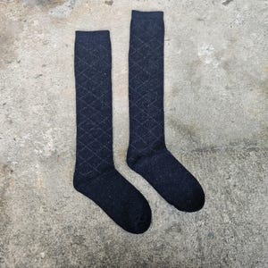 May include: A pair of dark navy blue knee-high socks. The socks feature a diamond pattern in a lighter shade of blue. The socks are made of a soft, possibly knitted material. The socks are displayed on a concrete surface.