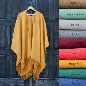 Puede incluir: Un poncho de lana amarillo mostaza con flecos. El poncho está colgado en una puerta de madera.