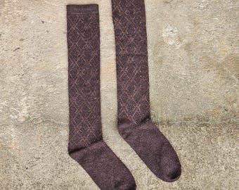 May include: A pair of knee-high socks in a dark brown color with a diamond pattern. The socks are made of a soft material and are designed to be worn on the legs. The socks are laying on a concrete surface.