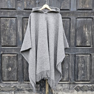 K&ouml;nnte beinhalten: Ein grauer Kapuzenponcho mit Fransen am Saum h&auml;ngt an einem Holzb&uuml;gel. Der Poncho besteht aus einem dicken, gewebten Material. Der Hintergrund ist eine dunkle Holzt&uuml;r.