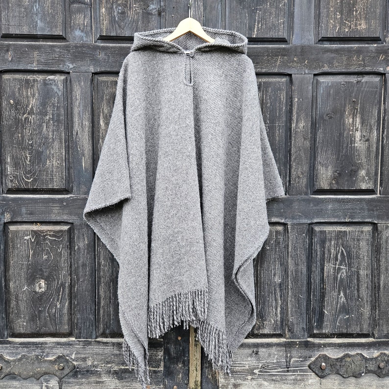 K&ouml;nnte beinhalten: Ein grauer Kapuzenponcho mit Fransen am Saum h&auml;ngt an einem Holzb&uuml;gel. Der Poncho besteht aus einem dicken, gewebten Material. Der Hintergrund ist eine dunkle Holzt&uuml;r.
