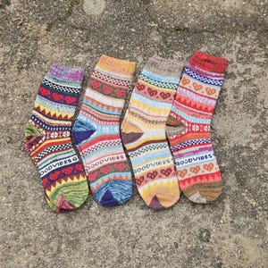 May include: Four pairs of knit socks featuring colorful horizontal bands with heart patterns, geometric shapes, and the text GOODVIBES printed in white. Each sock displays a unique combination of red, blue, yellow, green, and tan yarn colors.