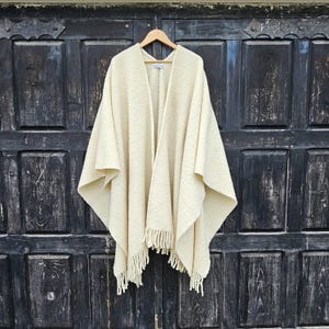 Op de afbeelding: Een crèmekleurige gebreide poncho met franjes. De poncho hangt aan een houten deur.