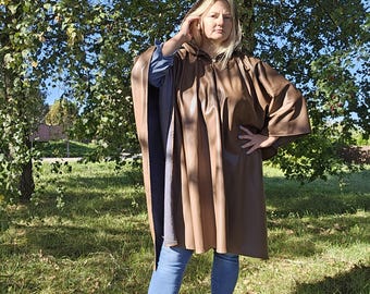 May include: A brown leather hooded poncho with a zip fastening. The poncho is being worn by a person with long brown hair and is standing in a grassy area.