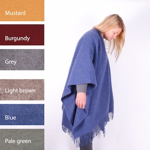 May include: A blue wool poncho with fringe. The poncho is shown on a model. The model is wearing the poncho over a grey long-sleeved shirt and dark jeans.